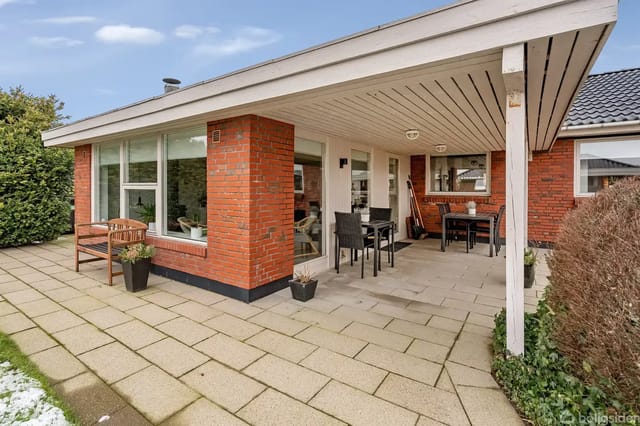 A red brick house on a paved path, surrounded by lawn with patches of snow. Several windows and a white door are visible, surrounded by bare shrubs.