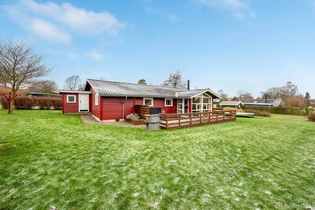 A red wooden cottage stands quietly on a green lawn, surrounded by trees and a clear blue sky. The cottage has large windows and a wooden terrace.