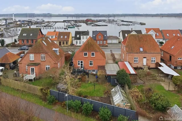 Red brick houses with red roofs and gardens along a street, with a marina and boats in the background.