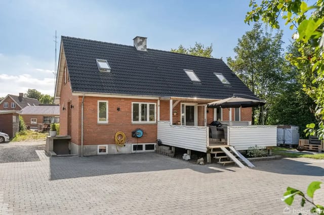 House with red brick and black roof, featuring a raised terrace with parasol and stairs, set in a driveway surrounded by trees and other houses in the background.