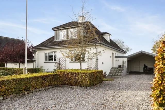 White house with dark roof in a well-maintained garden. Gravel path leads to a carport. Hedges and trees surround the area under a clear blue sky.