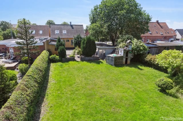 Green garden surrounded by bushes and trees, with a well-maintained lawn, seen from a high perspective. Background with red brick houses and blue sky.