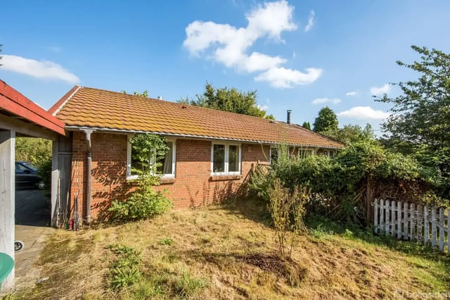 A small, brick-clad bungalow with a red tile roof stands quietly in an overgrown garden surrounded by trees and a white picket fence. Photographed in sunlight against a blue, cloudless sky.