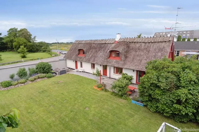 House with thatched roof and red windows, surrounded by well-kept lawn and bushes, located in a rural area with road and fields in the background.