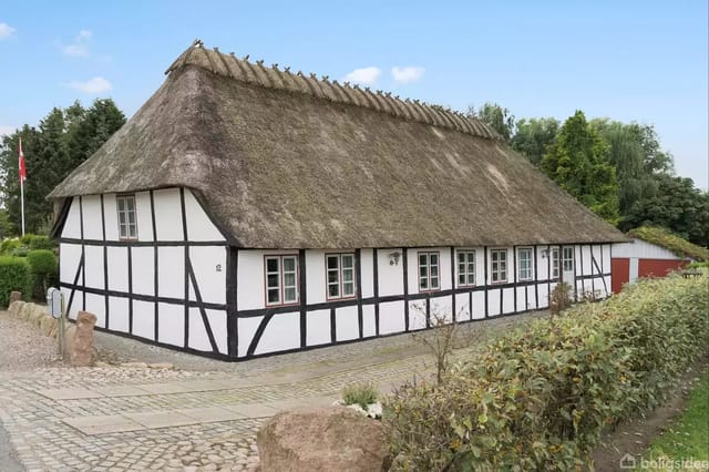Traditional timber-framed house with thatched roof on a cobblestone road, surrounded by hedges and trees in a rural setting.
