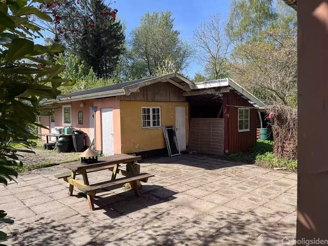 A wooden cabin stands quietly surrounded by bushes and trees in a sunny courtyard with a picnic bench in the foreground.