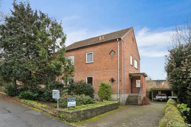 A red brick house in a quiet residential neighborhood with a 'For Sale' sign and a driveway leading to a carport.