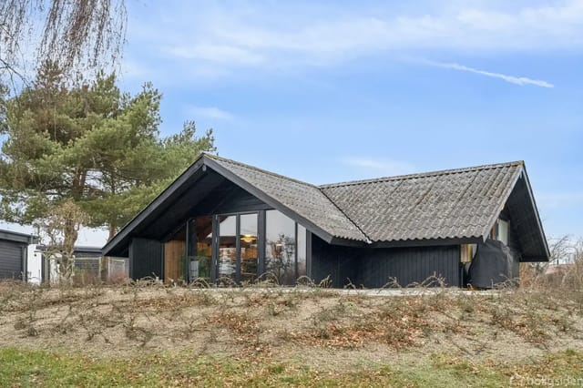 A black-painted house with large windows stands on a grassy hill surrounded by trees and a clear blue sky.