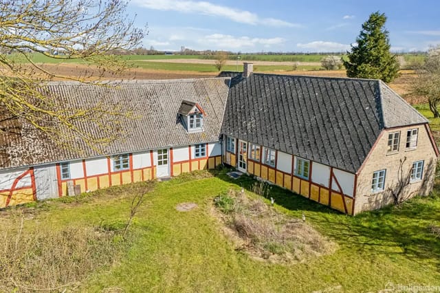 Half-timbered house with red cross patterns standing on a green lawn surrounded by open fields and trees under a blue sky.