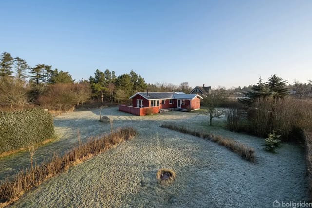 Red house stands quietly in a lush garden under a clear blue sky. Surrounded by trees and bushes, it creates a peaceful, rural atmosphere.