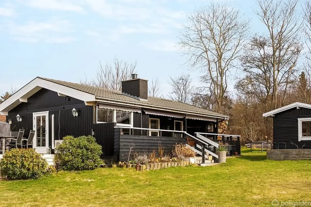 A black-painted wooden house with a large terrace surrounded by a green garden. A smaller outbuilding stands to the right. The background contains leafless trees.