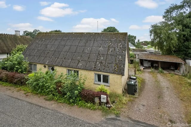 Yellow brick house with a slightly worn appearance, located by the road and surrounded by grass and bushes. A gravel driveway leads to a carport.