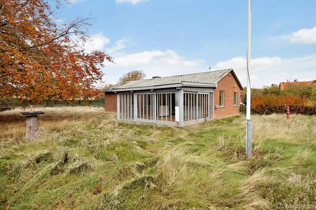 A red brick house with an integrated glass conservatory, surrounded by tall grass and bushes under a blue sky with scattered white clouds. A flagpole is placed in the foreground.