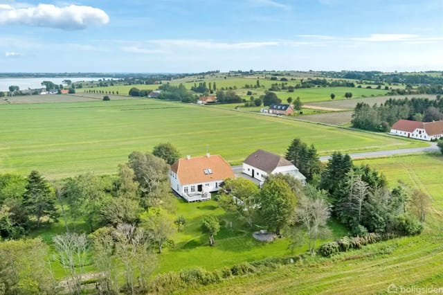 A white villa with a red roof stands among trees and bushes in a rural area with green fields and scattered houses nearby. A fjord is visible in the background.