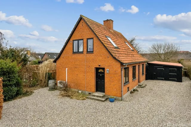 Red brick house with steep roof and skylights, located by a gravel courtyard with a black garage in the background. The house is surrounded by bushes and trees.