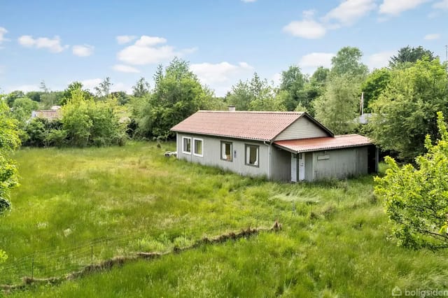 A gray-painted, simple house with a red roof stands quietly in an overgrown garden surrounded by tall grass and trees, under a clear blue sky with scattered clouds.