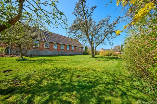 A long, red brick building stands quietly in a green garden with large, shady trees under a clear blue sky.