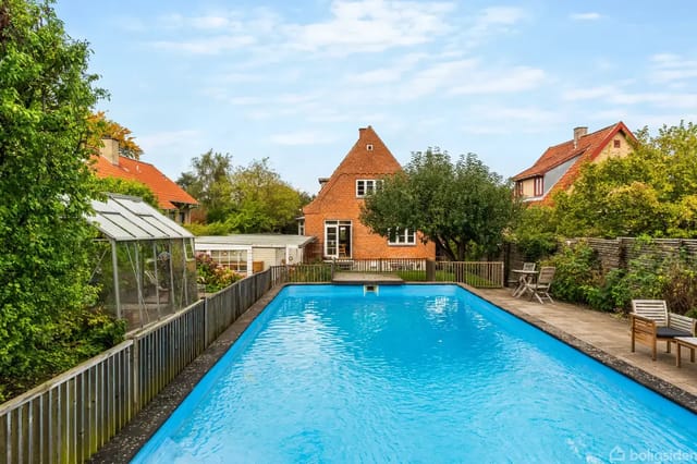 A rectangular swimming pool surrounded by a terrace with sun loungers. In the background, a red brick house with a garden of trees and bushes.