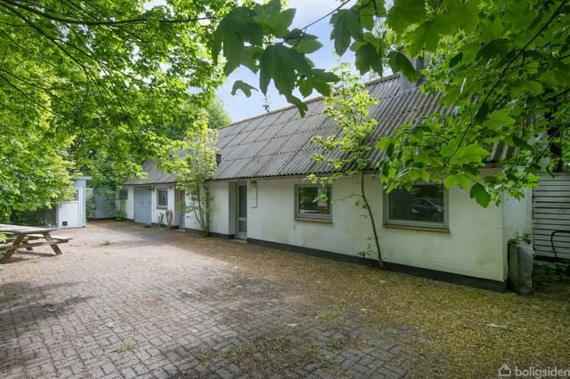 A white house with a sloped roof surrounded by green trees and a cobblestone courtyard.