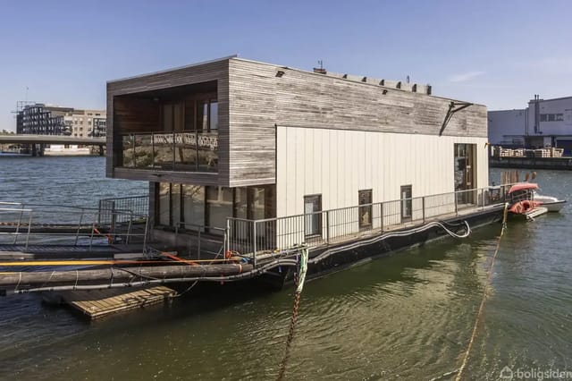 A modern houseboat moored at a quay, constructed of wood with large windows, floating on water in a city harbor with buildings in the background.