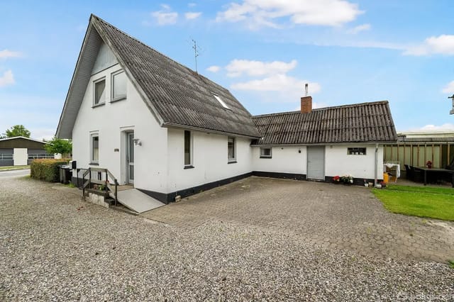 White house with sloped roof on a gravel lot, surrounded by a small lawn and clear sky.