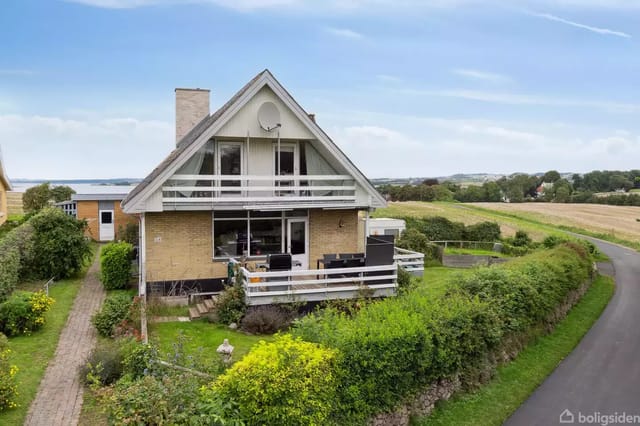 House with pointed roof and balcony among green bushes near a narrow road. The surrounding landscape shows fields and a view of the water in the background.