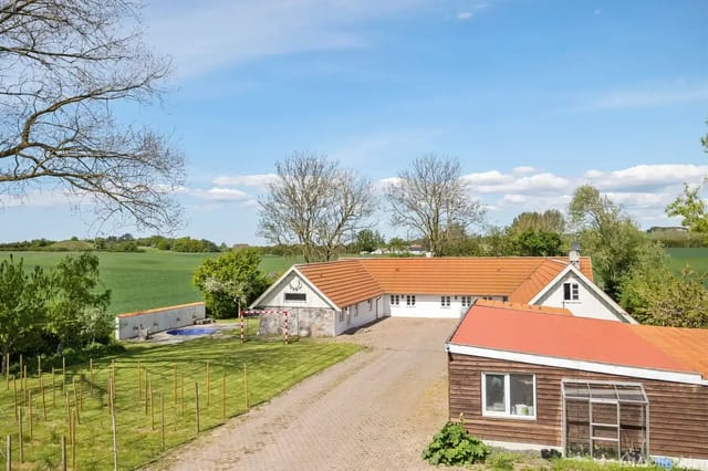 A white country house with an orange roof stands quietly surrounded by green fields and a few trees under a blue sky with clouds. A courtyard and driveway are visible in front of the building.