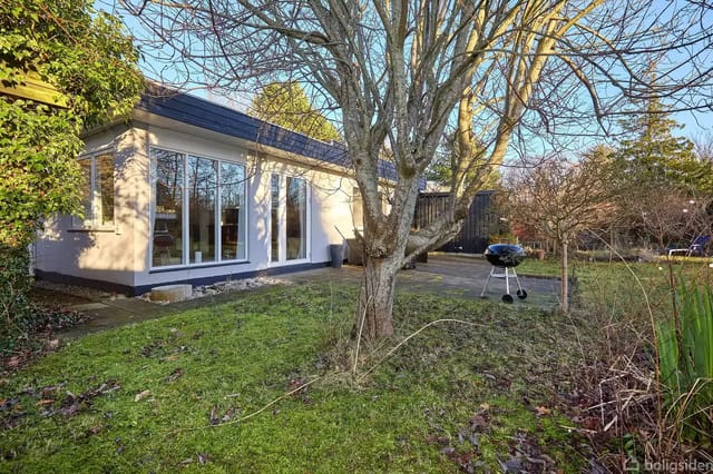 A house with large windows stands in a garden with a terrace. A grill is placed on the terrace, and grass surrounds the area with a central tree.