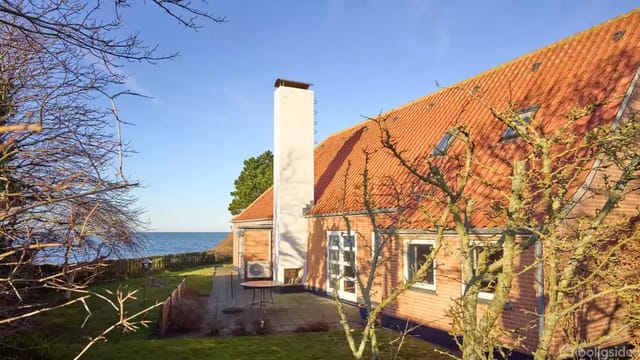 A brick house with a red tile roof and a white chimney stands on a plot with a lawn. Sea view in the background, surrounded by trees under a clear blue sky.