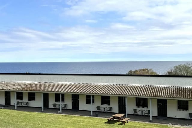 Building with flat roof on a lawn by the coast; the sea is visible in the background under a partly cloudy sky.