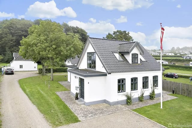 A white house with dark roof tiles stands on a terrace surrounded by grass. There is a Danish flagpole to the right, a driveway with a car to the left, and a forest in the background.