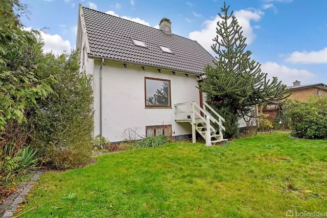 A white house with a sloped roof and dormer windows stands in a lush garden. A wooden staircase leads to a door. The garden surrounds the house with grass and bushes.