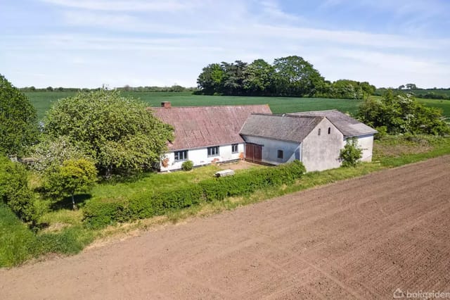 An old farmhouse with a red tile roof stands on a large field, surrounded by green trees and a light blue sky.