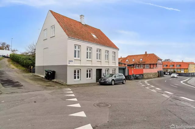 A white two-story house with a red tiled roof stands at the corner of a street. A car is parked in front of the house. The context is an urban street with several homes.