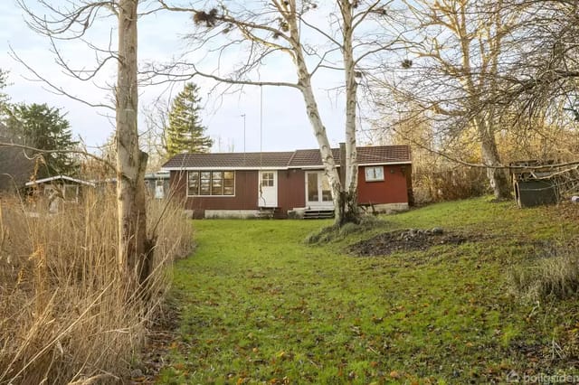 A small red house in a green garden surrounded by tall trees. Dark roof and white window frames are visible. The grass is green and tree branches stretch over the house.