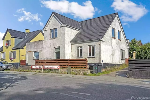 A white two-story house stands by a road with a sign in front indicating "Køge 20" and "Store Heddinge 10". The neighboring house is yellow with flowers in the windows.