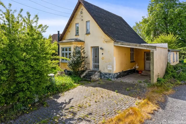 Yellow house with black roof, partially covered by green bushes, located by a cobblestone courtyard with overgrown vegetation.
