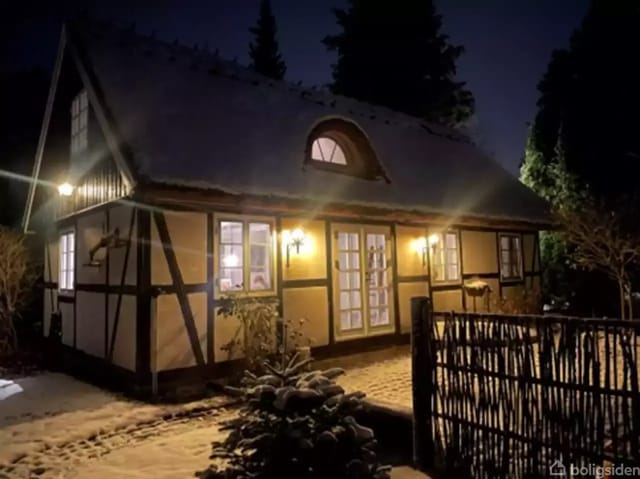 A timber-framed house with illuminated windows stands in a snow-covered garden at night. Snowy paths and trees surround the house, highlighted by outdoor lamps.