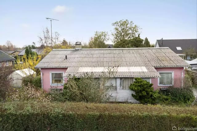 A small, pink house with a gray wave asbestos-free roof surrounded by bushes and trees in a suburb. Neighboring houses and a blue sky are visible in the background.