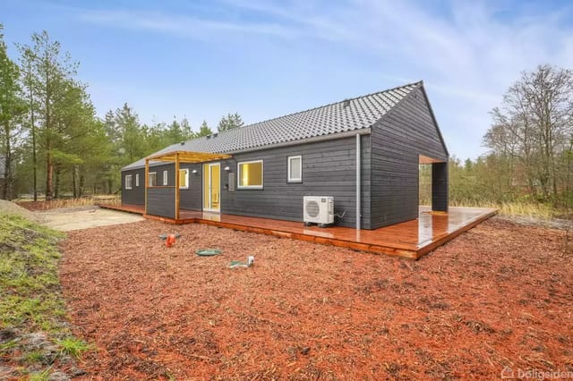 A black-painted wooden house with a gray tile roof on a bare plot, surrounded by forest.