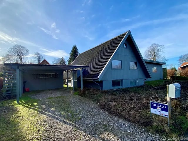 A-shaped house in a sunny garden with a steep black roof and blue walls, carport and 'For Sale' sign in the foreground.