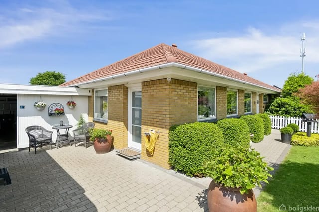 A brick house with a red tile roof, surrounded by a well-kept garden with shrubs and potted plants. There are garden furniture and a wall decoration by a paved terrace.