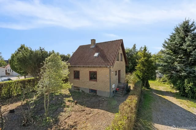 A brick house stands quietly surrounded by a garden with trees and bushes. A gravel driveway leads up to the house, which has a red tiled roof and a chimney.