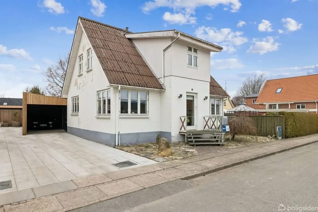 A white house with a brown roof in a residential neighborhood. There is a carport and a driveway to the left. The sky is clear and blue.