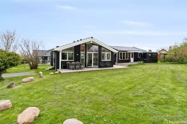 Black house with large windows surrounded by a well-kept lawn and bushes. A terrace in front of the house's glass facade is furnished with garden furniture.