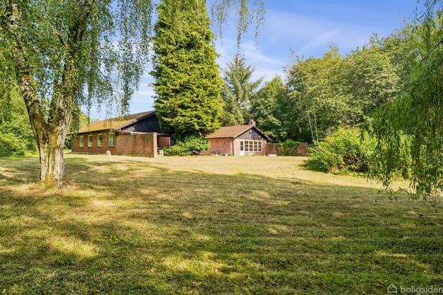 A brick house stands quietly in a wooded garden with well-kept grass and scattered trees.