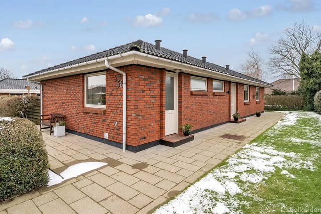 A red brick house on a paved path, surrounded by lawn with patches of snow. Several windows and a white door are visible, surrounded by bare shrubs.