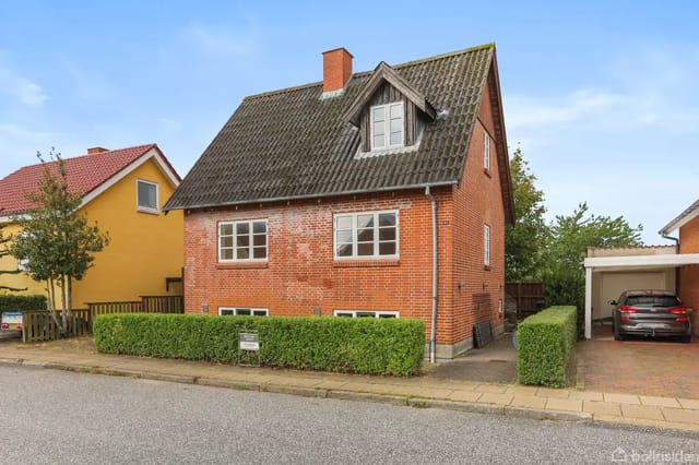 Red brick house with steep roof and dormer, located on a quiet residential street. Gravel driveway leads to a carport with a car. Green hedges surround the property.