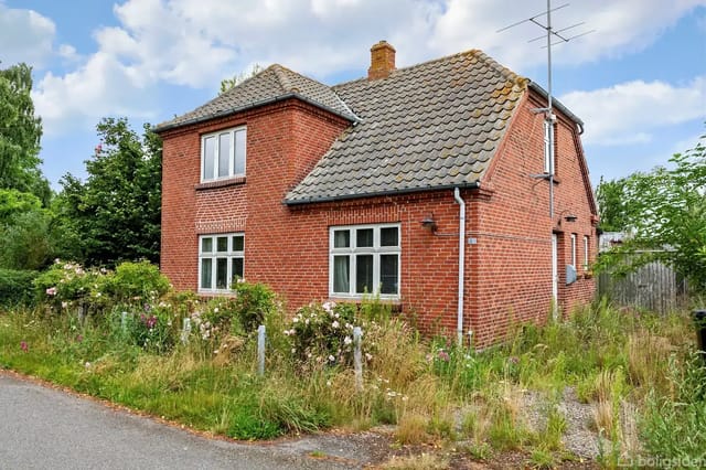 A red brick house with a gray tiled roof stands on an overgrown plot by a country road. The house's windows have white frames. An antenna is mounted on the roof.