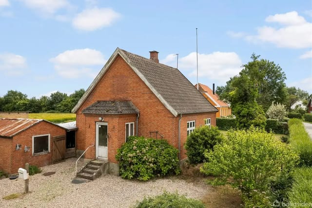 A red brick house with a steep roof pitch stands quietly next to a gravel driveway. To the left is a low outbuilding, surrounded by green bushes and trees.